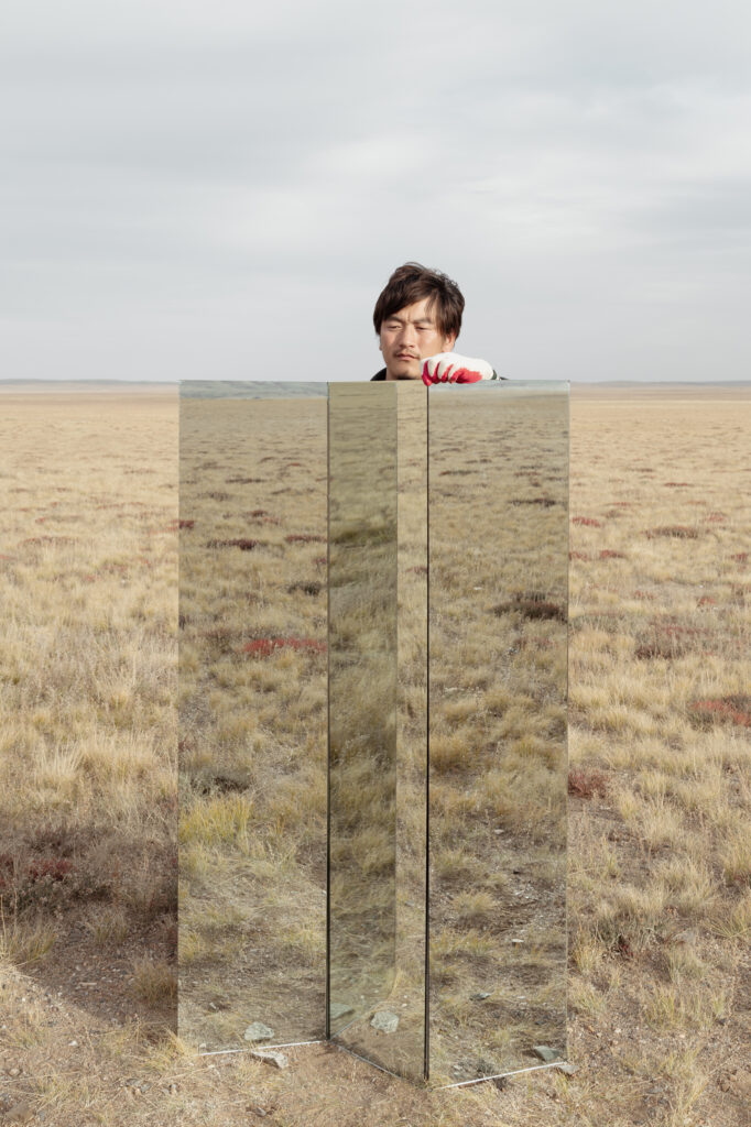 Eine kleine gerahmte fotografische Arbeit, auf der die mongolische Steppenlandschaft abgebildet ist. im Vordergrund steht eine Installation aus großen rechteckigen Spiegel. Darüber schaut der Kopf eines mongolischen Mannes hoch.
